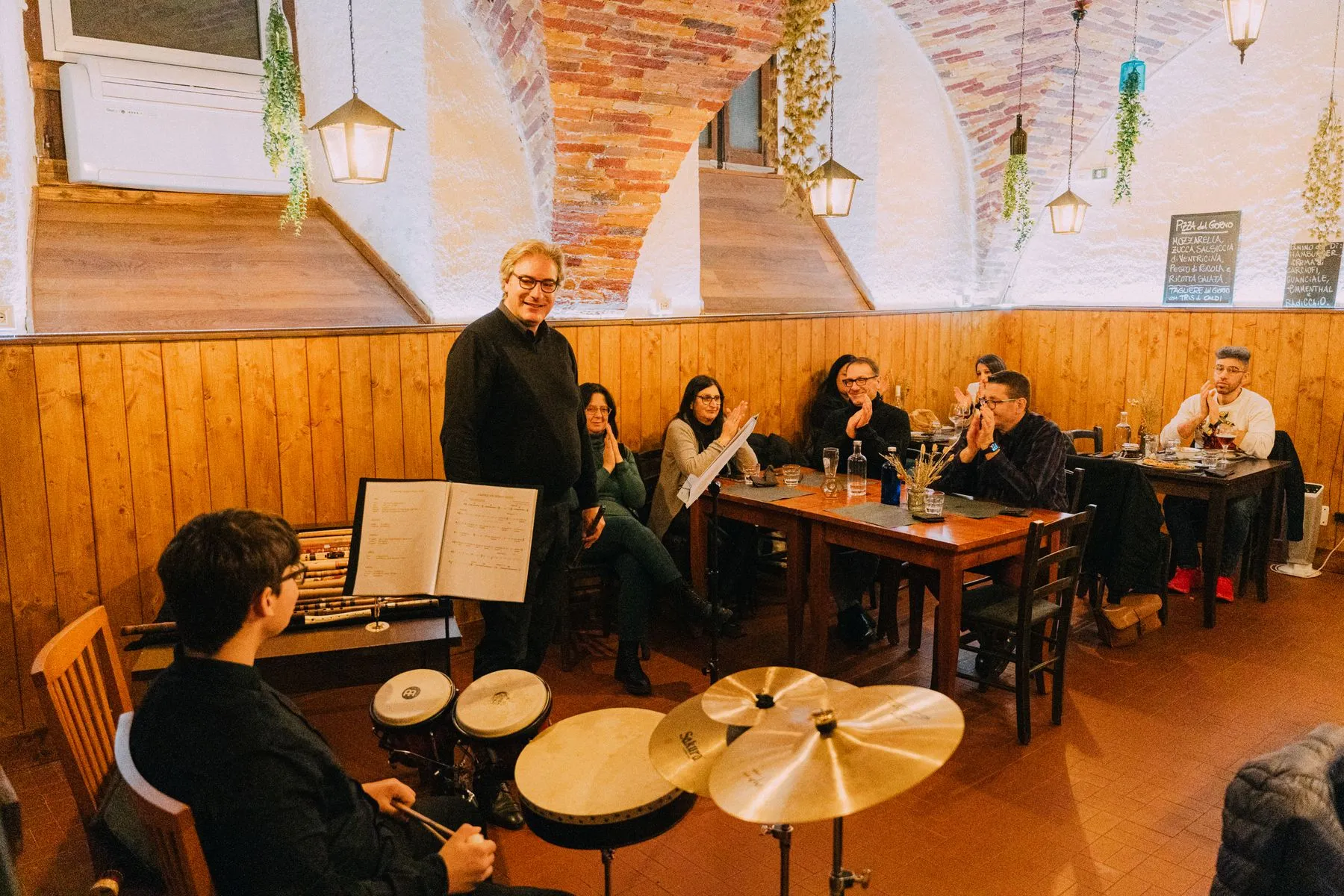 La sala durante la serata: il pubblico ai tavoli, le percussioni e il maestro che racconta
